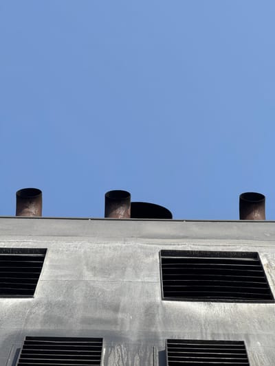 This media file captures a close-up, low-angle view of the side of a utilitarian concrete structure, possibly a ventilation tower or a component of an industrial building. The sky is a clear, bright blue, suggesting daytime and fair weather.
The structure itself is predominantly grey concrete, showing some signs of weathering or texture. Protruding from the side of the building are several cylindrical pipes, likely vents or exhaust ports. These pipes appear to be made of metal and have a rusty, weathered appearance.
In contrast to the open pipes, there are also several rectangular openings covered with black metal grates. These grates are composed of vertical bars and are set into the concrete surface, suggesting they are for ventilation or protection.
The composition is abstract, focusing on the geometric shapes and textures of the building and its components against the vast expanse of the sky. There is no indication of human presence or specific activity taking place. The lighting suggests a sunny day, casting subtle shadows that highlight the textures of the concrete and the edges of the grates and pipes. The overall impression is one of industrial architecture and functionality. The location context of Lorca, Spain, does not provide any direct visual cues within the image itself. No text is visible in the media file.