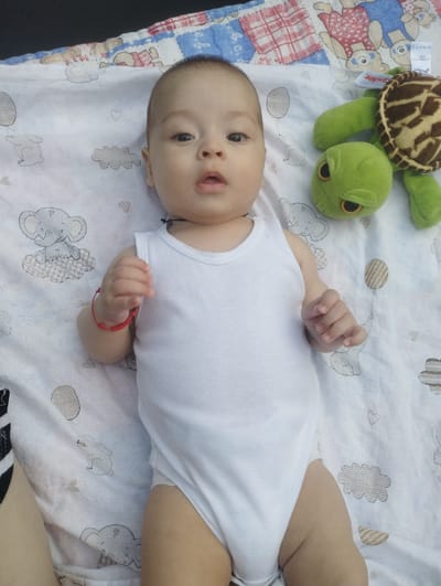 This is a medium shot, taken from above, of a baby lying on its back on a white blanket patterned with cartoon elephants, rabbits, and clouds. The baby is wearing a white tank-top style onesie. A plush green turtle toy is to the baby's right. A woven blanket with a red and white checkered pattern is visible at the top of the frame. The baby has a red bracelet on its left wrist. The lighting suggests it is daytime. The overall scene is calm and peaceful.