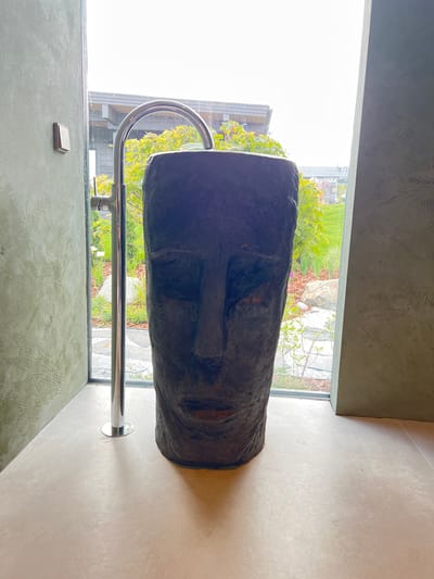 This image depicts a bathroom with a unique, face-shaped basin. The basin is dark gray and appears to be made of a rough material. A chrome faucet is installed behind the basin, arching over it. The bathroom floor is light-colored and appears to be polished concrete. Large windows overlook an outdoor area with greenery and some buildings in the distance. The walls are textured, with a green, patterned finish on the left and a more muted, textured finish on the right. There is a light switch on the wall to the left of the window. This scene is likely located in Yakunino, Russia, given the context provided.