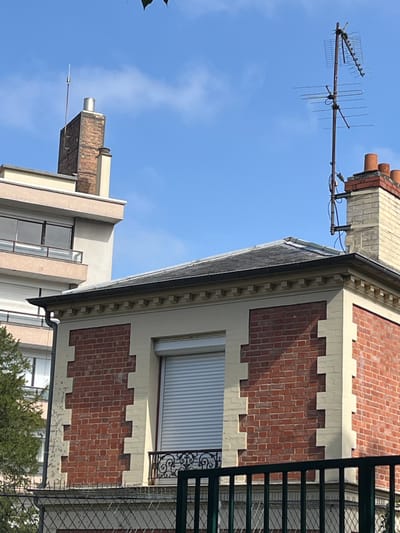 This image captures an exterior view of a two-story building in L'Haÿ-les-Roses, France. The lower story is constructed of red brick with decorative cream-colored accents. It features a large window with a white rolling shutter and a small, ornate black metal balcony railing. The upper story of this building is also cream-colored, topped with a dark gray roof. Adjacent to this building, to its left, is a taller, lighter-colored building with a balcony on its upper level. On the rooftop of the red brick building, a television antenna is mounted. To the left, a chimney made of darker brick rises above a lighter, rectangular structure. The sky is a clear blue with a few wispy clouds. In the foreground, a dark green metal fence is visible.