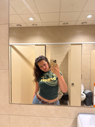 A woman is taking a selfie in a bathroom mirror. She is wearing a green t-shirt and jeans with a brown belt. The bathroom has tiled walls and a tiled ceiling with recessed lighting. There is a toilet and a sink in the background. The location is most likely not in Hadzhidimitrovo, Bulgaria, as there is no clear indication of the location.