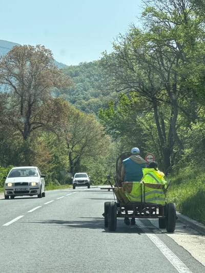 A road in Bilka, Bulgaria, shows a horse-drawn wagon occupied by two individuals wearing safety vests, followed by two cars. The wagon, appearing handmade, occupies the far right lane next to a road divider, suggesting it's sharing the lane with traffic. The cars follow behind, with a white sedan in the foreground. The surrounding environment includes trees and a distant mountain.