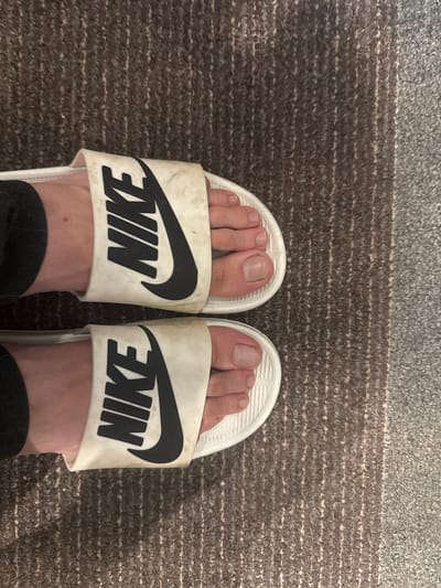 The image shows a close-up view of a person's feet wearing white Nike slides with black Nike logos. The slides appear worn and slightly dirty. The feet are positioned on a textured carpet with brown and grey stripes. The background is out of focus. There are no visible landmarks or indications of the specific location in Демихово, Russia.