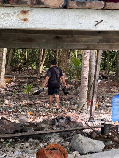 A man wearing a black t-shirt and shorts walks away from the camera under a wooden overhang. He is carrying a black pan in his right hand and a black object, possibly a shoe, in his left hand. He is in a wooded area with many palm trees and debris on the ground, including coconuts and plastic bottles. The ground appears to be sandy and rocky. The location is Ujongsedheun, Indonesia.