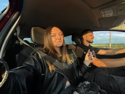 The image captures the interior of a car, seemingly from a wide-angle lens. A young woman with light brown hair, wearing a black leather jacket and a seatbelt, is facing the camera. Beside her, a man with a beard and a black t-shirt is visible, smiling. They appear to be inside the car. Through the car windows, a landscape with trees and sky is visible. The interior car details, such as the roof, seats, and control panel, are also visible. Considering that the location in the city of Babovo, Bulgaria, is provided, the background landscape could likely be in that region.