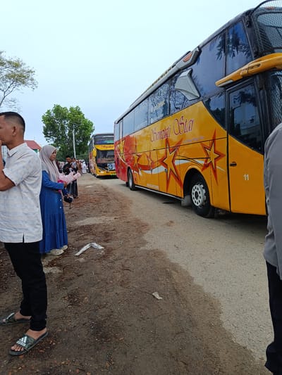 The image depicts a street scene in Meureudu, Indonesia, where several yellow and orange buses are parked. The foreground shows the lower bodies of three individuals, two men and one woman. The woman, on the left, is wearing a blue dress and a light-colored hijab. The man to her right is wearing a white patterned shirt and black pants. His feet are visible wearing camouflage slide sandals with the Adidas logo. The man furthest right is partially visible, wearing a gray shirt and black pants.

In the background, a large yellow bus with "Sempati Star" written on its side is prominently featured, adorned with star graphics. Another similar bus is visible behind it, and a third bus, partially obscured, is further down the line. The ground appears to be a dirt and gravel road. The sky is overcast. There are trees and a building with a red roof visible in the distance. The scene suggests a location where people are either boarding or disembarking from buses, possibly a bus terminal or a pick-up point.