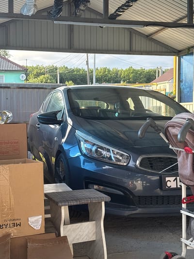 The image shows a dark blue Kia Ceed parked under a metal canopy. The car is dusty and has a license plate with the characters "C 15" visible. A baby stroller with a brown and beige patterned fabric is partially obstructing the view of the car's front. In the foreground, there are several cardboard boxes, one of which has "METAR" printed on it. There is also a small, two-tiered stool with a checkered fabric top and a marble-patterned base. The background shows a residential area with houses and trees. The location is identified as Sovkhoza "Razdol'e", Russia. The scene appears to be an outdoor private parking area or a yard.