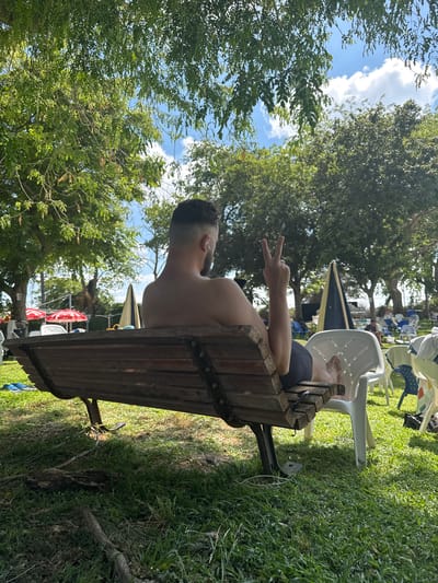 A man is sitting on a wooden park bench in what appears to be a resort or public park area, possibly in Beit Yitzhak-Sha'ar Hefer, Israel. He is facing away from the camera, wearing dark shorts. His left arm is raised, with his index and middle fingers extended in a peace sign gesture. Behind him, the environment consists of lush green grass, mature trees with dappled sunlight filtering through their leaves, and a bright blue sky with scattered white clouds. In the background, there are several white plastic chairs, some colorful beach umbrellas, and other park amenities. The overall atmosphere suggests a relaxed, outdoor setting.