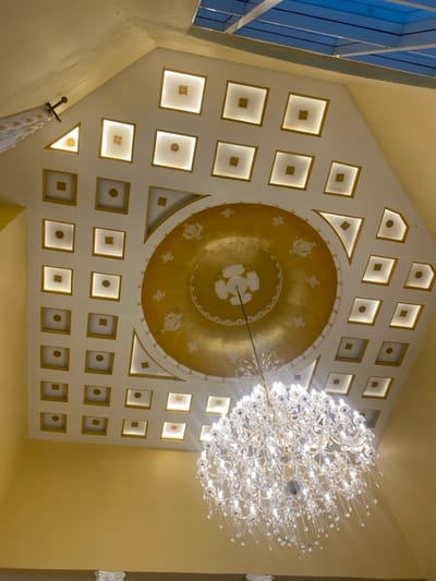 The image shows a ceiling in a building in Buea, Cameroon, located at the coordinates 4.145 latitude and 9.23 longitude. The ceiling has a white and gold color scheme, with a large, circular, golden dome at the center. The dome features intricate, faded, gold design patterns and is surrounded by a grid of 24 square, white panels. The panels contain smaller, square, gold shapes in their centers, each with a white light. A large crystal chandelier is hanging from the ceiling, with a thin, black chord connecting it to the dome. The chandelier is directly below the dome, positioned at the center of the grid of panels. The chandelier shines brightly, casting a warm light across the space. The building's walls are painted a soft yellow. The image captures a high-quality, detailed view of the ceiling. The image is most likely taken from the bottom, looking upwards towards the ceiling.