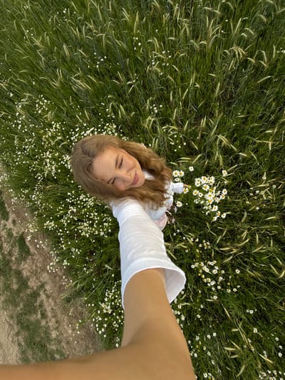 An overhead shot captures a young woman lying in a field of grass and daisies. She is looking upwards with her eyes closed and a gentle smile on her face. Her long, wavy blonde hair is spread around her. She is wearing a white, long-sleeved shirt. Her right arm is extended towards the camera, holding a bouquet of daisies. The field is dense with green grass and interspersed with numerous white daisies. The ground to the left of the image appears to be a dirt path or a less vegetated area. The scene is set in a natural outdoor environment, possibly a meadow or a field on the outskirts of the city of Snovitsy, Russia.