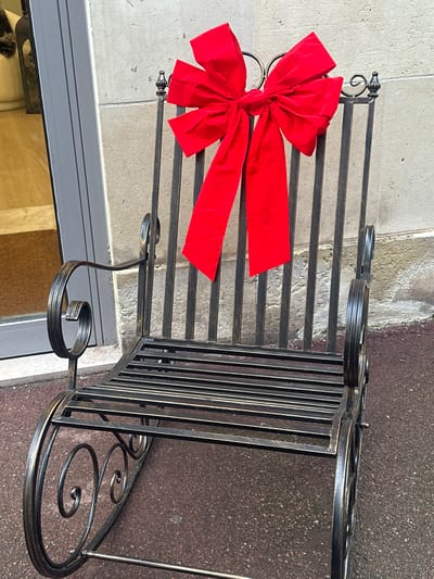 A dark, wrought-iron rocking chair, featuring intricate scrollwork on its armrests and rockers, is positioned outdoors on a reddish-brown textured pavement. The chair's backrest consists of vertical slats, and the seat is formed by horizontal slats. A large, bright red velvet bow is affixed to the upper portion of the chair's back. The background features a light grey stone or concrete building wall to the right and a dark grey door or window frame to the left. A light-colored object, possibly a display item, is partially visible inside the frame on the left, above a brown floor or mat. Sunlight from the left casts distinct shadows of the chair and bow onto the building wall. This scene is located in the city of Saint-Germain-en-Laye, France.
