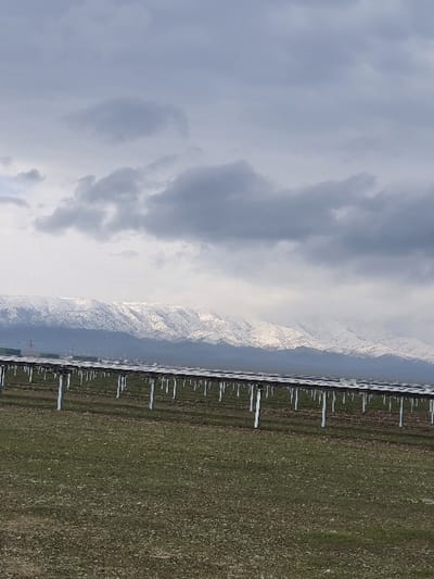 The landscape features a field of green dotted with repetitive white structures, likely solar panels, extending into the distance. Jagged, snow-capped mountains rise in the backdrop, partially obscured by haze. Overcast skies dominate the scene, with dense gray clouds filling the frame.