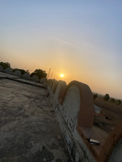 The image depicts a panoramic view of Barako, Nigeria, at either sunrise or sunset, characterized by a bright yellow sun low on the horizon. The foreground consists of a textured, dark grey concrete surface, possibly a rooftop. A decorative wall or parapet runs diagonally across the midground, featuring light grey blocks with reddish-brown arched coping stones. On one of these coping stones, a dark electronic device, likely a smartphone or power bank, is visible with a white cable attached. A small, light brown rectangular object rests on the concrete surface near the wall. In the background, silhouetted against the warm, gradient sky (light blue transitioning to yellow-orange), are trees, several tall communication towers to the left of the sun, and an open, sparsely vegetated landscape to the right, which includes a distant small, light-colored building. No individuals are present.