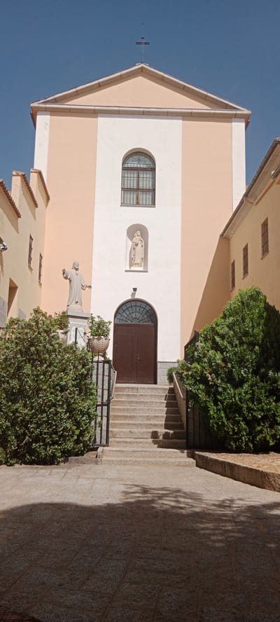 The image shows the facade of a church, likely the parish church of San Lorenzo de El Escorial, Spain. The building has a pale peach facade with a central white vertical section. At the top of the facade, a cross is visible against a clear blue sky. Below the cross, there is a large arched window with a decorative grille. Beneath this window, in a recessed niche, stands a statue of a robed figure holding a book. Further down, another arched window or decorative element is above a dark brown double door. Flanking the entrance are stone stairs leading up to the door. To the left of the stairs, a statue of a standing male figure with outstretched arms is mounted on a pedestal. Lush green bushes and trees are planted on either side of the entrance and in pots near the statue. The ground in front of the church is paved with large stone slabs, and shadows from unseen foliage are cast upon it. Adjacent buildings, painted in a similar pale peach and yellow hue, are visible on the left and right sides of the main church structure, with arched windows and sloped roofs. A black metal railing runs along the side of the stairs and the bushes. The overall impression is of a sunny day in a town square or church courtyard.