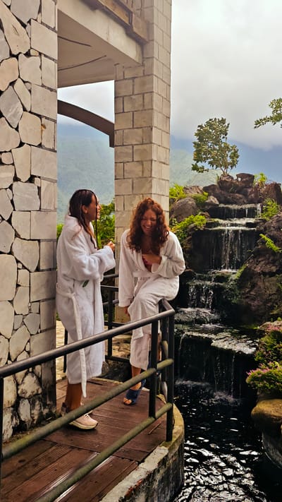 Two women wearing white bathrobes stand on a wooden platform enclosed by a black metal railing in Cat Cat Village, Vietnam. The woman on the left has dark hair and is facing the woman on the right, who has red curly hair and is perched on the railing. A stone structure flanks them on the left and a brick structure is behind the woman on the right. A waterfall cascades down rocks and greenery to a dark pool of water. Behind the waterfall, mountains are visible in the distance. The woman on the left wears tan sneakers, while the woman on the right wears a blue sandal.
