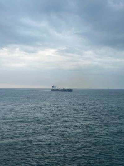 A long cargo ship is sailing on a choppy, dark blue ocean under a cloudy, grey sky. The ship is in the middle distance, moving from left to right. Several other smaller cargo ships are visible on the horizon, further in the distance. The sky is overcast with light and dark grey clouds, obscuring any direct sunlight. The water is a deep blue-grey with small waves. The overall scene suggests a possibly cool or overcast day at sea. The location is unknown, Malaysia.