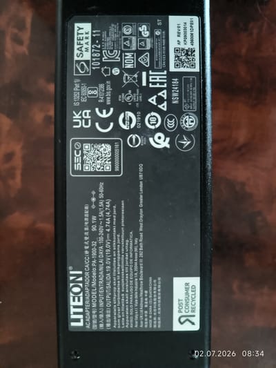 The image is a close-up, top-down shot of a black power adapter. The adapter has various labels with technical specifications and certifications. There are no people, the environment, or any elements related to the city of Kattasu, Uzbekistan present in the image. The focus is solely on the product's labeling.