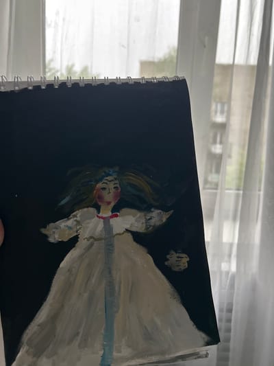 The image shows a spiral-bound sketchbook lying against a window. The sketchbook is open, displaying a painting of a figure with pale skin, painted in white and pale tones. The figure has a red face and neck, light brown hair, and a blue headpiece. The figure is wearing a white gown and is positioned against a solid black background. Behind the sketchbook, there is a window with horizontal blinds. The background shows an indistinct view of buildings and greenery.
There is no indication of the city Dmitrievskaya in Russia.