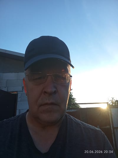 An outdoor, medium-shot, selfie captures a Caucasian man wearing a dark blue baseball cap and glasses. He has fair skin and appears to be in his late 50s to early 60s. He is wearing a dark grey t-shirt. The man is looking directly at the camera. The background consists of a bright, clear blue sky with the sun setting on the right side of the frame. The sun's rays are visible, creating a lens flare effect. To the left of the man, a portion of a building made of grey cinder blocks is visible. The man is positioned in what appears to be a rural or suburban outdoor setting, potentially in or around the city of Урыцкі сельскі Савет, Belarus, given the provided context. The image is dated 20.06.2026 at 20:34.