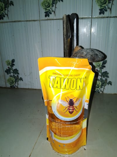 The image depicts a close-up of a yellow pouch of "TAWON" brand cooking oil, branded as "Minyak Goreng Sawit". The pouch is positioned vertically and features a prominent illustration of a bee. Behind the pouch, a dark, vaguely wedge-shaped object is visible, possibly a tool or a piece of agricultural equipment, resting against a tiled wall. The wall is adorned with a repeating pattern of stylized green roses. The overall setting appears to be an indoor space, likely a kitchen or storage area, given the tiles and the presence of household items. The floor is tiled and appears somewhat dirty with scattered dark specks. The lighting is dim, creating shadows. The image does not contain any people or specific identifiable landmarks of Sendangrejo, Indonesia, but the product branding suggests a connection to the Indonesian market.