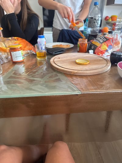 The scene depicts an indoor domestic setting in Kanchevo, Bulgaria, likely a dining or kitchen area, centered around a wooden table covered with a clear plastic protector. Three individuals are partially visible. In the foreground, the bare legs and lower thighs of an individual are visible beneath the table. To the mid-left, a person with long, dark hair, wearing a black long-sleeved top, is seated with their left hand raised towards their mouth/face. To the mid-right, a person wearing a white short-sleeved top is actively preparing food, holding a piece of bread with their left hand and using an orange-handled knife in their right hand to spread a red condiment from a small glass jar onto the bread.
The table is laden with various items:
On the left, an open orange plastic bag contains bread rolls. Next to it, a glass jar with a yellow lid and a label (partially visible) stands beside a small glass containing an amber liquid. A white plastic bottle with a blue cap is also present.
Further right, a black bi-fold wallet rests near a white paper plate holding a piece of bread with red spread. A smaller glass jar containing red condiment is next to it.
In the center-right, a large clear plastic bottle of dark liquid (Coca-Cola brand visible) stands beside a bag of potato chips with yellow and red packaging ("КУЛИНАР ПАРТИ" visible). A stack of clear plastic cups and another clear plastic bottle containing water are also on the table. A pink and white bowl is partially visible on the far right.
A round wooden cutting board occupies the central area of the table, with a yellow metal jar lid placed flat on its surface.
In the background, a white shelf or cabinet unit is visible, along with a bowl containing assorted fruit. The floor is light-colored laminate.