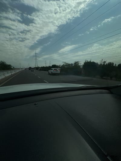 The image shows a view from inside a vehicle traveling on a highway in Istiqlol, Uzbekistan. The sky is partially cloudy with a mix of white and gray clouds against a blue sky. Several power lines stretch horizontally across the frame, supported by tall lattice towers visible in the distance. The highway has multiple lanes, with other vehicles on the road. One car, a white SUV, is visible in the right lane. On the left side of the highway, there is a concrete barrier and then a strip of vegetation. On the right side, there is a dense line of trees and bushes. The foreground of the image is dominated by the dashboard and windshield of the vehicle from which the photo was taken.