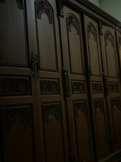The image shows a close-up of a dark wooden wardrobe with intricate carvings on its doors. The wardrobe appears to be large, with multiple doors visible in the frame. Each door has a distinct design, featuring arched panels with detailed floral and geometric patterns carved into the wood. The handles are also ornate, made of dark metal. The lighting in the image is dim, casting shadows that highlight the texture and depth of the carvings. The overall impression is of a traditional and well-crafted piece of furniture. The location in Manado, Indonesia, is not evident from the image itself.