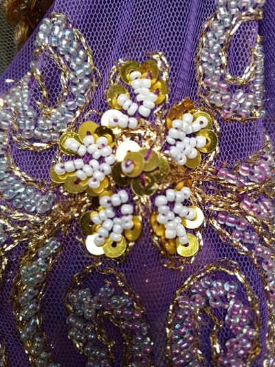 This close-up image captures a section of intricately embroidered purple mesh fabric, likely from a traditional garment or decorative item in Houaykhang, Laos. The fabric serves as a delicate base for a rich array of embellishments.
A prominent central motif features a five-petaled flower, where each petal is composed of clusters of opaque white seed beads surrounding individual, bright gold sequins. These floral elements are meticulously stitched onto the purple netting.
Extending from this central design and across the fabric are elaborate swirling patterns and abstract shapes, precisely outlined with shimmering gold metallic thread. Within these outlines, sections are filled with small, translucent iridescent seed beads. These beads exhibit a beautiful color play, shifting between light blue and purplish-pink hues, adding depth and sparkle to the overall design.
The fine, open weave of the purple mesh is clearly visible beneath the dense beadwork and sequins, providing a striking contrast to the opulent embellishments. The detailed craftsmanship highlights the artistry involved in creating this ornate textile. No specific activity is depicted, and no text is visible.