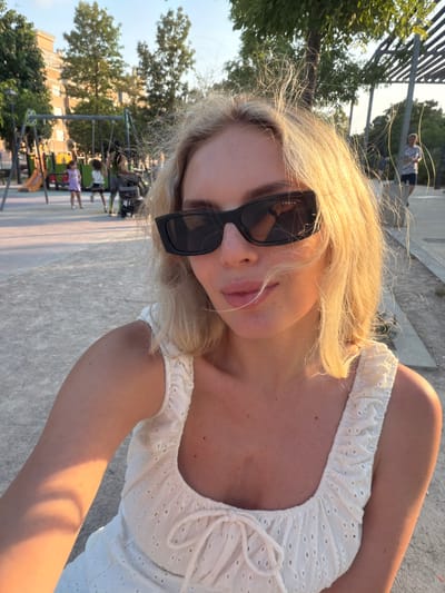 The image shows a close-up selfie of a young woman with blonde hair and sunglasses, standing in an outdoor public space. She is wearing a white, eyelet-fabric top with ruffled straps and a drawstring tie at the bust. The background reveals a playground with swings and slides, children playing, and trees. In the distance, there are buildings and a pedestrian walkway with modern, slatted overhead structures. The lighting suggests it is late afternoon or early evening, with warm sunlight casting shadows. The setting appears to be a park or recreational area within a residential urban environment. Based on the provided information, the location is identified as Benimàmet-beniferri, Spain.