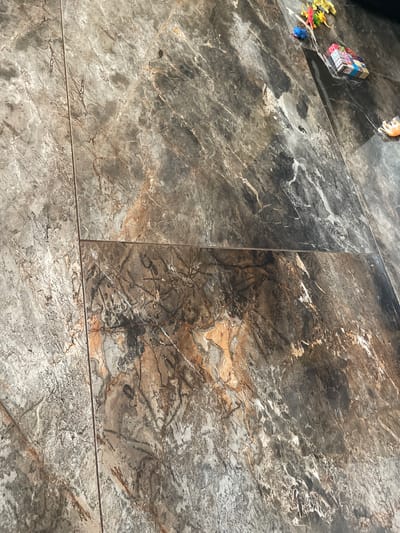 The image shows a close-up view of a marble-patterned floor. The floor tiles are large and have a dark gray and brown color scheme with white and orange veining. There are some small toys scattered on the floor in the upper right portion of the image, including toy dinosaurs and a blue toy creature. The lighting suggests an indoor environment. There is no specific indication of the location in Yasen, Bulgaria, beyond it being a floor.