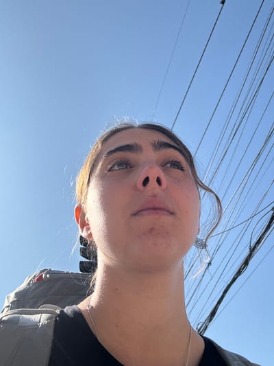 An upward-angled selfie features a young woman under a bright blue sky cluttered with power lines. She is fair-skinned, with prominent eyebrows and light-colored eyes. There are some blemishes on her cheeks. A silver necklace adorns her neck, and a small earring dangles near her hair, which is pulled back. A glimpse of her clothing suggests a dark top with a gray jacket or backpack. The sun casts a strong light on her face, creating shadows and highlights.