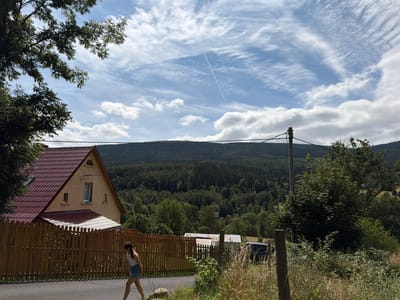 An outdoor, medium shot captures a woman walking on a paved road adjacent to a wooden fence. The woman, positioned in the lower left of the frame, wears a white tank top and denim shorts, with her body angled away from the camera. Behind the fence, a yellow house with a red tiled roof is visible. The background features a dense forest that extends to a distant, tree-covered mountain range under a sky with scattered clouds and a faint airplane contrail. The scene is set in Świeradów-Zdrój, Poland, suggesting a rural or semi-rural environment with a clear day.