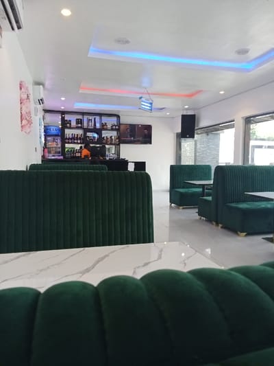The image shows the interior of a restaurant or bar, located at 5.471, 5.818 in Ujevwu, Nigeria. The space has a modern, sleek design with white walls, a white tiled floor, and a white marble countertop. There are several green velvet booths with high backrests arranged around the space. The booths are well-maintained with the upholstery appearing clean and new. The walls are adorned with a few decorative elements. A large, wooden bookshelf filled with bottles and other items is on the left side of the image. The shelves are lit from below. A flat-screen television is mounted on the wall to the right of the shelves. The space appears quiet and clean. The only visible person is a figure with an orange shirt standing in front of the shelves, but their face is not visible. The scene is empty, lacking any action, interaction, or people.