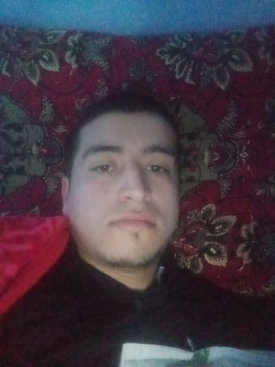 An adult male is depicted in a close-up, frontal view, likely a selfie, within an indoor environment. He possesses dark hair and facial hair in the form of stubble or a short goatee, with a neutral expression directed towards the camera. He is wearing a dark-colored garment, possibly a long-sleeved shirt or hoodie, with a portion of lighter fabric visible on the lower right of the frame, potentially part of his clothing or an object in front of him. To the immediate left of the subject, a section of plain red fabric, possibly a blanket or cushion, is visible. The background consists primarily of a dark red patterned textile, possibly a rug or tapestry, featuring floral or ornamental designs in shades of green, black, and beige. A narrow strip of a plain, light blue surface, likely a wall or ceiling, is visible along the top edge of the image. The lighting appears dim and possibly artificial, contributing to a somewhat grainy quality. The precise location within Kasan, Uzbekistan, cannot be determined from the visual evidence presented.