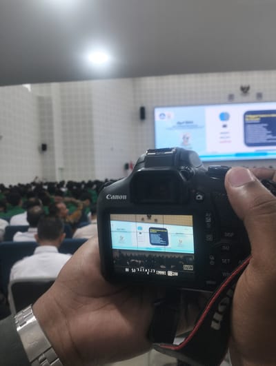 The image shows a person holding a Canon camera, with the camera's LCD screen displaying a live view. The camera is pointed towards a lecture hall where a crowd of people are seated, facing a large screen at the front. The screen displays a presentation with text and graphics. The environment appears to be indoors, with ceiling lights visible. The person's hands are visible holding the camera and a watch is worn on the left wrist. The camera settings displayed on the screen include "80 5.0 1/125 123 [9999] ISO 200". The presence of a large lecture hall and presentation screen suggests an academic or professional event is taking place. Given the context of a lecture hall, it is likely that this image was taken in a public or institutional building within the city of Reuleut Timu, Indonesia, possibly at an educational institution or conference center.