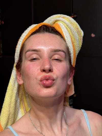 A close-up, rotated portrait captures a young person indoors, likely in Заболотово, Russia. The subject has a striped towel, featuring yellow, orange, and white bands, wrapped around their head, suggesting recently washed or wet hair. Their face appears damp and has a noticeable sheen, with areas of redness on the cheeks, indicating a recent facial treatment or skincare routine. The person's lips are pursed in a playful "kissing" or "duck face" expression, and their eyes are partially closed. A slender silver chain necklace adorns their neck, and a light blue bra strap is visible on their right shoulder. The background is dark and out of focus, with a faint, blurry heart-shaped light reflection visible in the upper left corner, typical of indoor bokeh. No visible text is present in the image. The overall impression is one of relaxation and playfulness after a self-care activity.