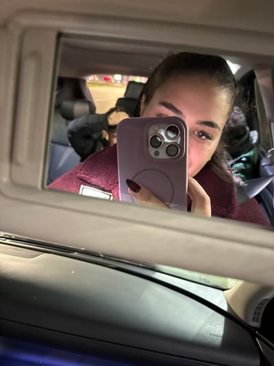 A selfie taken in a car mirror. The woman in the reflection is holding a purple iPhone, its camera lenses visible. Her eyes are visible above the phone, and her nails are painted a dark red. She wears a maroon-colored top with a patch. The car's interior is visible, including a black panel with "AIRBAG" printed on it. Through the mirror, the blurry outline of a person seated in the car and exterior lights can be seen.