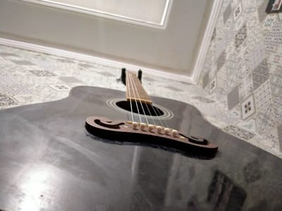 A black acoustic guitar is lying on its back, its strings and bridge facing the camera. The guitar is in a room with white walls and a tiled floor. The tiled floor is covered in geometric patterns in shades of grey, white, and black. The location is in the city of Lagadar, Indonesia at coordinates -6.928 latitude and 107.535 longitude. The camera is positioned close to the guitar, giving a wide-angle view of the instrument. There are no people present. The image is taken in a low-light environment, and the guitar's surface reflects some of the light. The room appears to be cluttered and potentially unfinished, with a white ceiling that is not fully finished.
