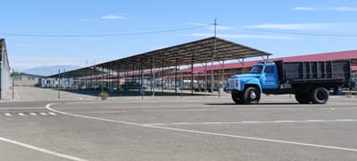 The scene shows an outdoor marketplace in Hovtashen, Armenia on a bright, sunny day. The marketplace consists of rows of metal-framed stalls covered by large awnings with a red roof, presumably to protect vendors and customers from the elements. An empty blue truck with a dark wooden cargo bed is parked in the foreground on a paved parking lot with white lane markings. The truck has Armenian text on the driver's side door and a number "5" visible on the front. A chain-link fence borders the stalls and parking area. A few smaller buildings are visible in the background, behind the fence. In the distant background, a range of mountains is visible. The sky is clear with scattered clouds.