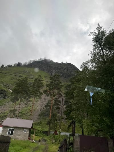 The image depicts a mountainous, rural landscape under a cloudy sky. In the foreground, a small, gray, rectangular building with a brown, corrugated metal roof is visible. It has at least two windows, one larger with a white frame and one smaller. A wooden porch extends from the front of the building. Adjacent to the house, there is dense green vegetation and trees, including several tall pine trees. A dirt path leads away from the house towards the viewer. In the mid-ground, a steep, grassy mountainside rises, with rocky outcrops and scattered trees. The upper slopes of the mountain are shrouded in mist. In the background, the mountain continues to rise, with its peak partially obscured by clouds. The overall impression is one of a remote and somewhat rugged environment. The provided location "Uznezya, Russia" suggests this image was taken in a rural area within that region. There are no people visible in the image.