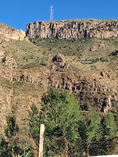 The image captures a steep, rocky mountainside covered in dry grass and sparse vegetation. A prominent cliff face with layered rock formations dominates the left side of the frame, with a towering electrical pylon visible in the distance on the summit. The right side of the image shows more of the grassy slope, with a cluster of green trees partially obscuring a wire fence and a weathered wooden post. The scene appears to be set in a dry, arid environment under a clear blue sky, suggesting a sunny day. The orientation of the image is rotated 90 degrees counter-clockwise. There is no visible text. The setting suggests a natural landscape, possibly a mountainous region in Shatin, Armenia, given the location context. The overall impression is one of rugged, untamed nature.