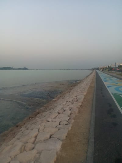 The image depicts a coastal scene in Al Khobar, Saudi Arabia, taken near sunset or sunrise. On the left is the sea, with a rocky embankment leading to the water. The water appears calm, with some sediment near the shore. On the right is a paved walkway with a painted lane designated for pedestrians and cyclists. People can be seen walking and cycling in the distance along the walkway. Streetlights line the road further down, with some cars driving. The sky is overcast, contributing to the subdued lighting. The overall scene suggests a recreational area by the sea.