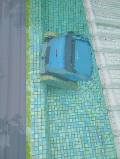 An aerial, high-angle, full shot shows a blue and gray automatic pool cleaner submerged in a residential swimming pool. The pool cleaner is positioned in the center of the frame, with its blue top lid and gray base clearly visible. The pool water is clear, with a tiled bottom that reflects the light in a mosaic pattern of green and blue squares. To the left of the pool cleaner is a white pool cover with subtle gray stripes. To the right of the pool cleaner are the white steps of the swimming pool, also submerged in water and exhibiting a textured surface. The overall scene suggests the pool is either being cleaned or has recently been cleaned. The scene takes place in Nowa Wieś Szlachecka, Poland.