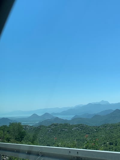 The image depicts a scenic mountainous landscape, likely in the vicinity of Gornja Brova, Montenegro. The foreground features a metallic guardrail, indicating a roadside viewpoint. Beyond the guardrail, lush green vegetation covers rolling hills and more distant, undulating mountains. The mountains recede into the background, becoming progressively hazy and blue-toned, suggesting atmospheric perspective. A body of water, possibly a lake or bay, is visible in the middle ground, nestled between the closer hills and the more distant mountain ranges. The sky is a clear, vibrant blue, with no clouds visible. The overall scene conveys a sense of expansive natural beauty on a bright, clear day. There are no people or animals present in the image.