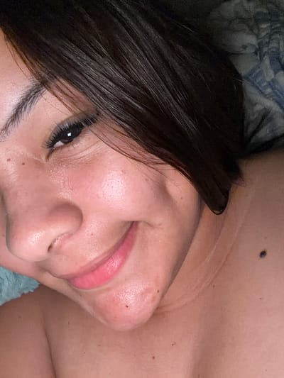 A close-up, upper-body portrait shows a woman with dark hair and fair skin smiling. She is lying down, with her head tilted slightly to the left. Her eyes are dark and wide, and her lips are painted a bright pink. Her hair is dark brown and falls in soft waves around her face. There is a slight sheen to her skin, suggesting it may be oily or that she is wearing makeup. A dark mole is visible on her neck, to the right of her face. The background is blurred and appears to be a patterned fabric, possibly a blanket or pillow. The image is lit from above, creating soft shadows on her face. The overall impression is one of relaxation and contentment. The image was taken in Balch Springs, United States of America.