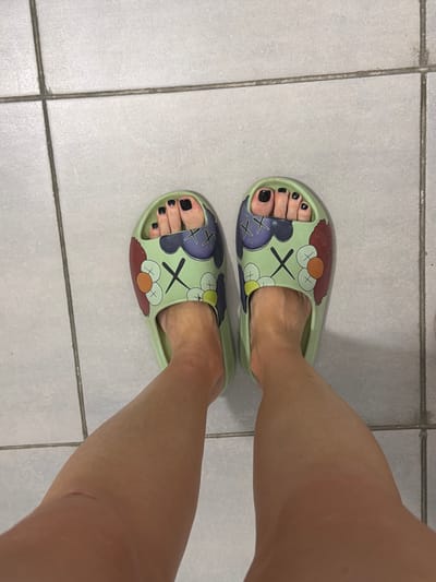 The image shows a pair of feet wearing light green slide sandals with a cartoonish design featuring "X" eyes and circular shapes. The toenails are painted black. The feet are standing on a tiled floor, with grout lines visible. The floor tiles are square and light gray. The lighting appears to be artificial. The image is a close-up, waist-down view of the subject. The location in Novoalexandrovka, Russia is not discernible from the visual information provided.