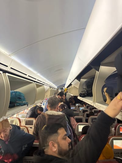 The image displays the interior of a commercial aircraft cabin, observed from the aisle, likely at Charles de Gaulle Airport in Tremblay-en-France, France. The scene is crowded with approximately 15-20 visible passengers, primarily adults, standing or moving in the aisle. White overhead bins line both sides of the cabin; several are open, revealing various pieces of luggage including a bright blue backpack on the left and a dark blue bag with a light-colored garment on the right.
In the foreground on the right, a man with a beard, dark hair, and an earring is captured in profile, facing left. His right arm is raised, possibly in the act of stowing or retrieving an item from an overhead bin. To his immediate left, another man with light brown hair, wearing a brown hooded top, is seen from behind. In the lower-left foreground, an older woman with light hair, wearing a dark blue top over a red garment, is partially visible. Further along the aisle, additional passengers are positioned between rows of reddish-brown upholstered seats, each equipped with an individual entertainment screen, some displaying illuminated content. The cabin's ceiling is white, featuring integrated lighting. The overall environment suggests a state of active passenger transit, consistent with boarding or deplaning operations.