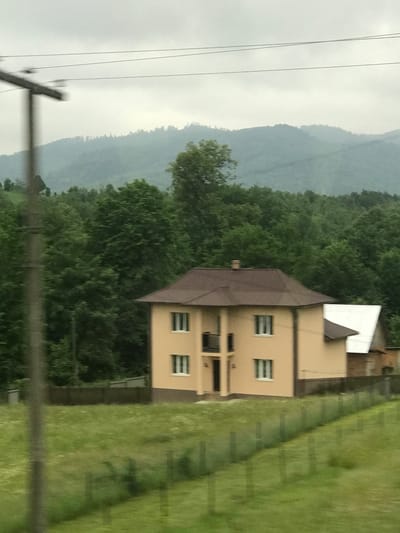 This image captures a rural scene in Krasnoilsk, Ukraine, under an overcast sky. In the foreground, a verdant green grassy field slopes upwards, partially divided by a wire fence running diagonally from the mid-left to the lower right. A blurred utility pole is prominently positioned on the far left. The midground features a two-story, light beige-colored house with a dark brown, multi-pitched roof. The house has multiple rectangular windows with dark frames and a small second-floor balcony with a dark railing centered above a dark main entrance. To the right of the primary residence, a smaller auxiliary building with a distinct white roof and darker-toned walls is partially visible. A dark fence encloses the property surrounding the houses. Immediately behind the buildings, a dense forest of various green deciduous trees stretches across the landscape. The background reveals multiple layers of tree-covered mountains, appearing muted bluish-grey due to atmospheric perspective. The sky is uniformly light grey and cloudy, with faint horizontal lines, likely power lines, traversing the upper portion near the utility pole. No people are present, and no specific actions are depicted.