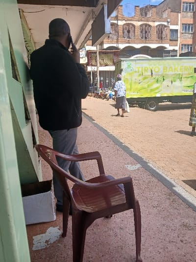 The image depicts an outdoor urban scene in Huruma ward, Kenya, during daylight. In the immediate foreground, a dark reddish-brown plastic armchair is positioned facing away from the viewer on a reddish-brown speckled pavement. To the left of the armchair, a white cardboard box rests against a light green vertical panel. A male individual, viewed from behind, stands partially obscured by the green panel and the armchair. He wears a black long-sleeved top and grey trousers, holding a black smartphone to his right ear as if engaged in a call.

Beyond the male subject, a reddish-brown paved street extends into the midground. A female individual, also viewed from behind, walks away from the camera on this street, carrying a white bag in her left hand. She wears a light-colored patterned top and dark lower garments. To her right, a light green commercial truck with "TIRYO BAKERY" printed in black on its side is visible, displaying a yellow and green banana-themed graphic.

In the background, multi-story buildings constructed from reddish-brown brick or painted masonry feature multiple windows, some equipped with decorative metal grilles. Overhead, a clear blue sky with minimal cloud cover is visible. The scene is illuminated by natural sunlight, casting minimal shadows.