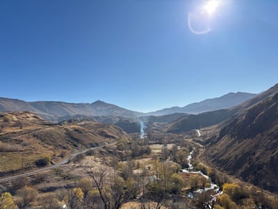 The image depicts a sunlit mountain landscape under a clear blue sky. A winding river flows through a valley dotted with trees, some bare and others with yellow leaves. A road traces along the side of the valley, leading towards the distant mountains. A plume of smoke rises in the valley. Hillsides transition from green to brown. Mountains fade into the hazy horizon.